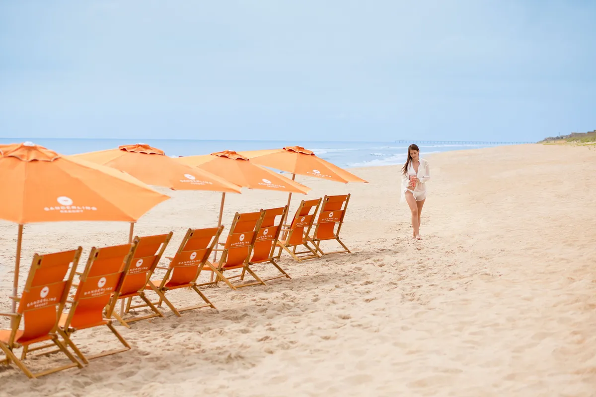 A cheerful private beach scene with beach chairs and umbrellas set up near the shoreline. A person is strolling along the sand, with gentle waves lapping in the background. The ocean and sky meet in a tranquil horizon, inviting guests to unwind, build sandcastles, or go for a refreshing swim.