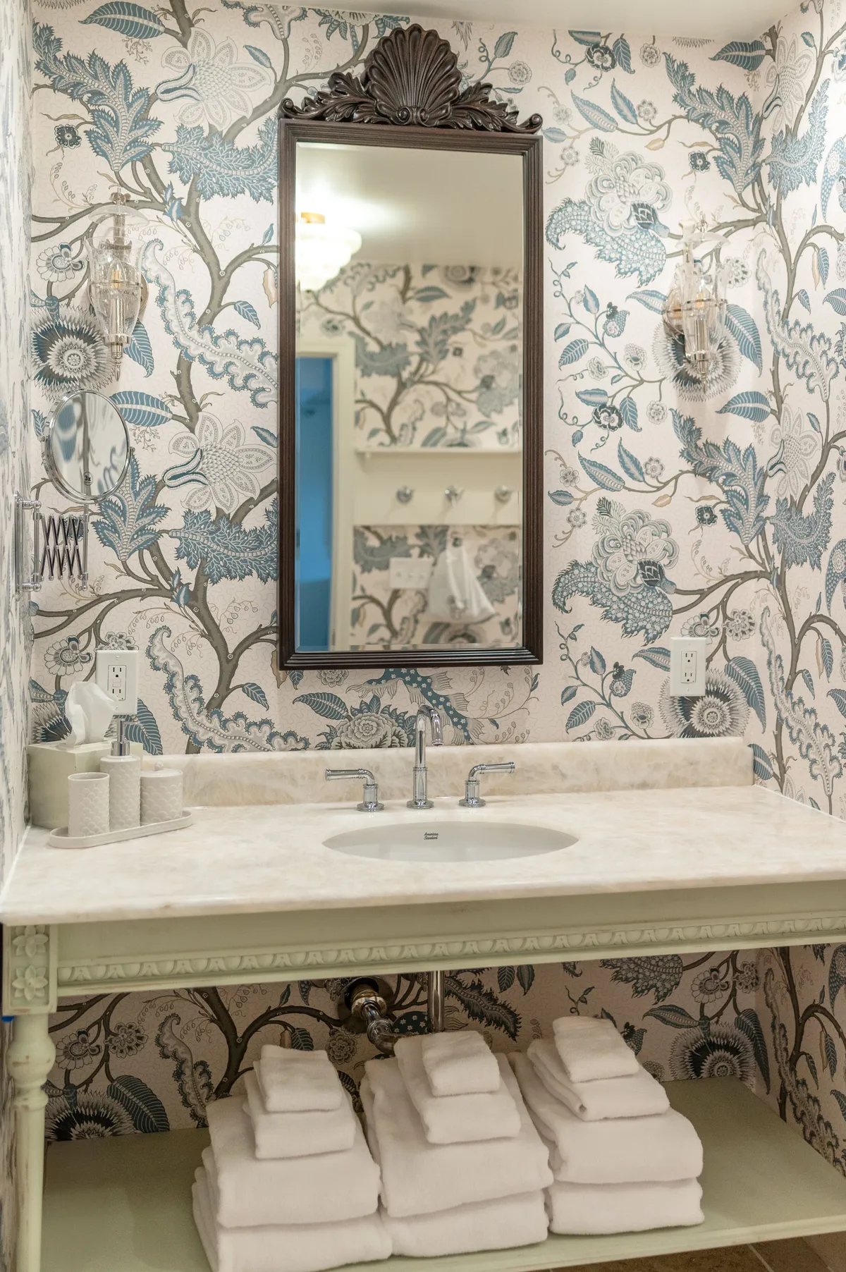 A beautifully appointed bathroom with an ornate mirror above a marble sink, set against elegant floral wallpaper. Neatly stacked white towels and soft lighting evoke a spa-like ambiance, inviting guests to indulge in self-care and relaxation.
