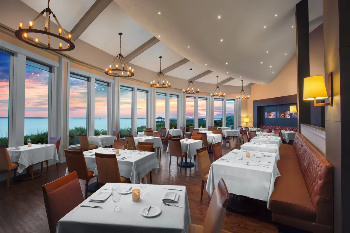 An upscale dining area with large windows offering ocean views, elegant chandeliers, and pristine white-tableclothed tables arranged with comfortable seating. The ambiance is sophisticated yet inviting, perfect for enjoying fresh, local cuisine in a beautiful setting.