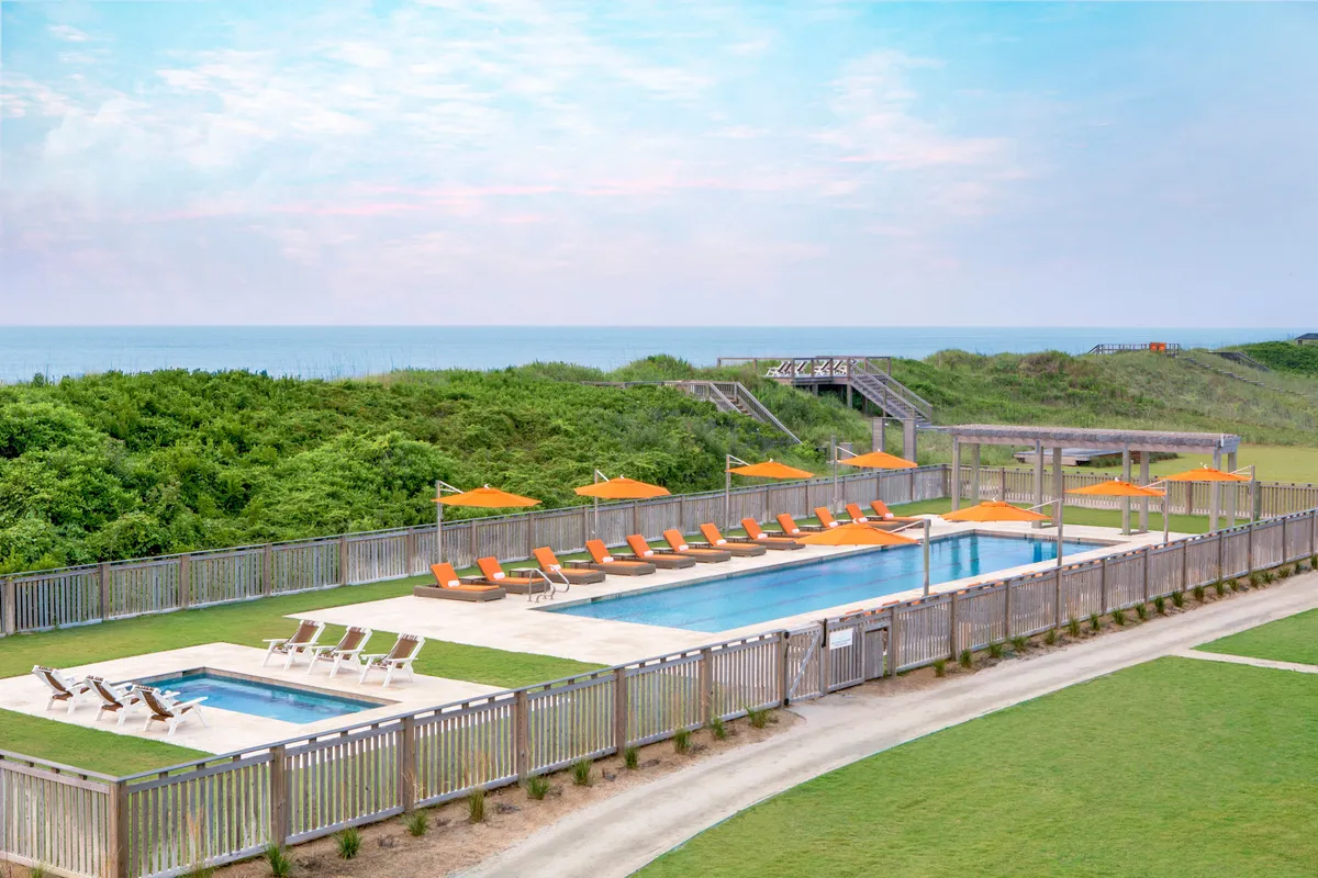 A vibrant outdoor pool scene with sparkling water, surrounded by inviting lounge chairs shaded by bright orange umbrellas. Lush green landscaping frames the pool area, while the ocean shimmers in the background. Several groups of people are relaxing, sunbathing, and enjoying the leisurely atmosphere, hinting at both quiet relaxation and lively social gatherings.