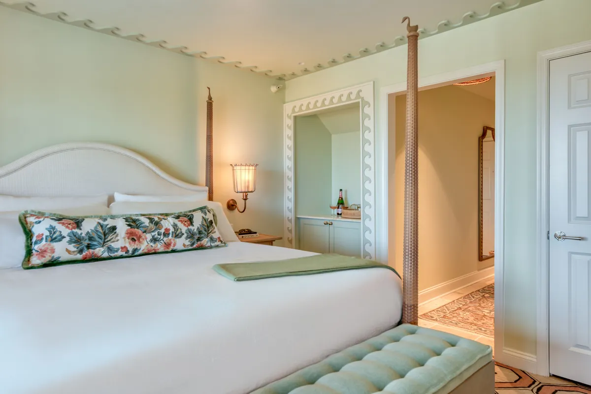 The image shows a serene studio interior featuring a plush king bed adorned with a floral pillow, a mirrored dresser that reflects soft natural light, and a decorative lamp. The setting is tranquil and inviting, suggesting relaxation and intimate moments, perfect for a couple. The overall ambiance is elegant yet comfortable, with understated, refined decor.