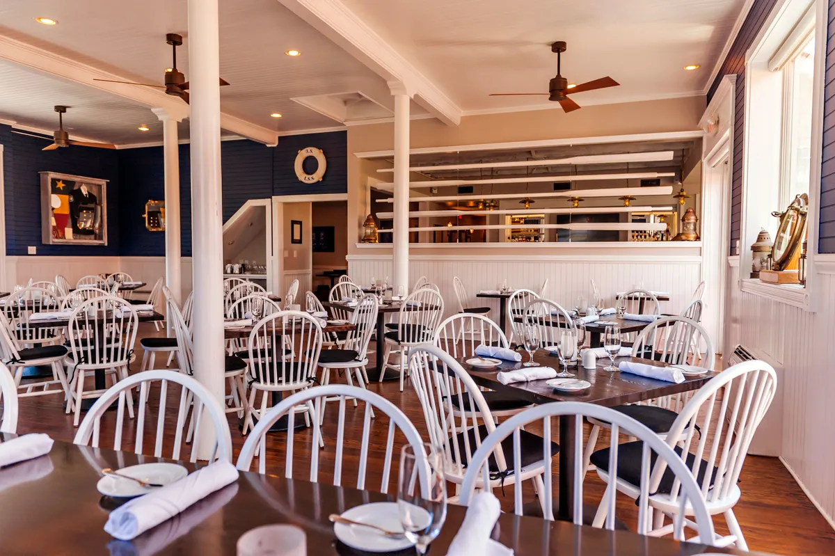 This image shows a welcoming interior dining space featuring wooden tables, white chairs, and a well-lit bar area. The atmosphere is relaxed, with people socializing and enjoying gourmet meals, hinting at a communal space where guests unwind and connect.