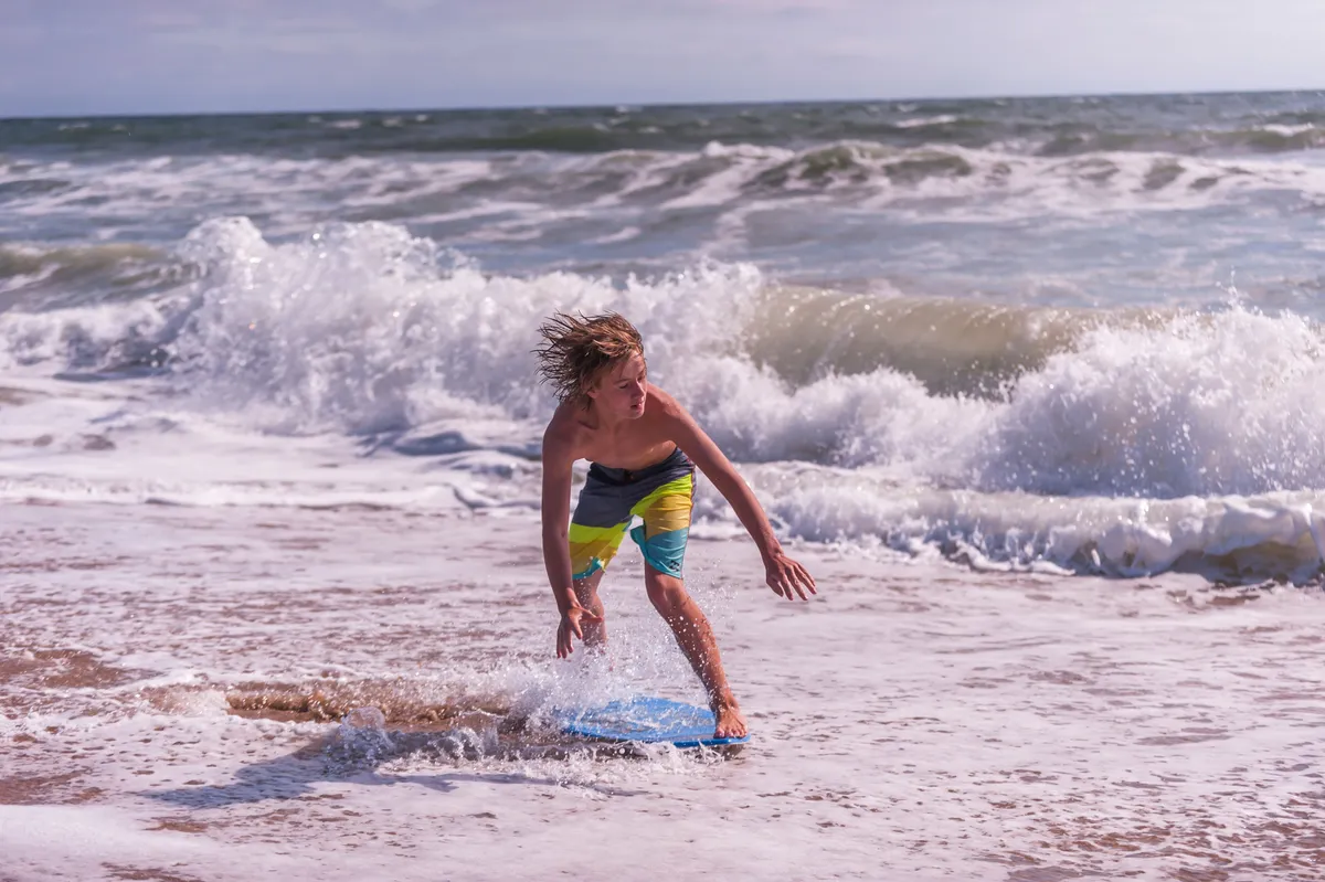 An energetic beach scene featuring a boy riding a boogie board as waves crash around him on the shoreline. The mood is playful and lively, with visible excitement and activity in a natural, sunlit setting.