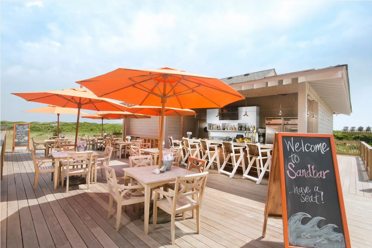 An inviting outdoor dining area with orange umbrellas, wooden tables, and a welcoming sign. The scene is casual and vibrant, set up for groups and families to enjoy meals together under the open sky. The area is bustling with guests savoring food and conversation, perfectly aligning with the description of on-site dining options.