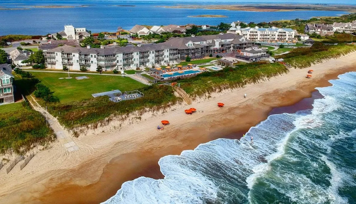 This image shows a stunning beachfront property with a swimming pool, beach umbrellas, and the ocean waves just beyond. The scene captures guests enjoying the sun on poolside loungers, strolling along the sandy shore, and the possibility of beach volleyball. The tranquil water and open sky suggest a classic Outer Banks vacation experience, ideal for families seeking both relaxation and recreation steps from their accommodations.