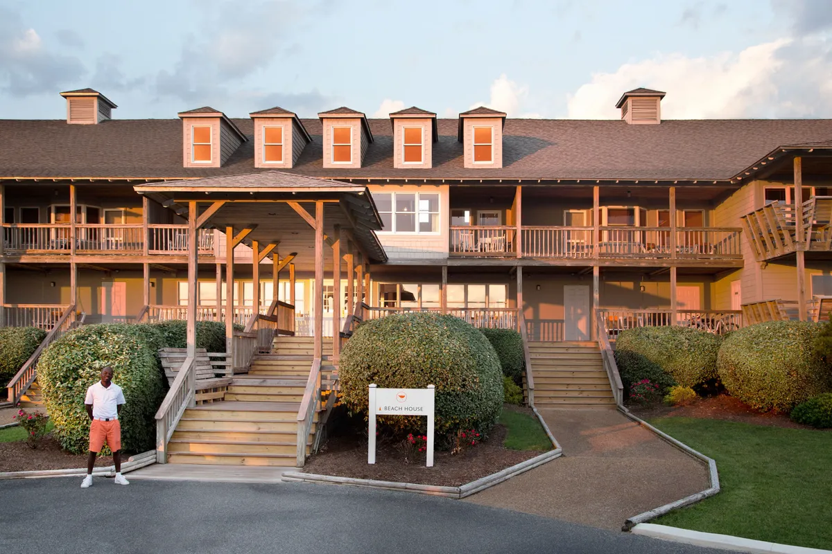 The inviting entrance to the beach house, framed by lush greenery, with a welcoming person standing at the forefront. The porch and deck hint at spaces for socializing, relaxing, or sharing meals, set against the backdrop of the nearby beach.