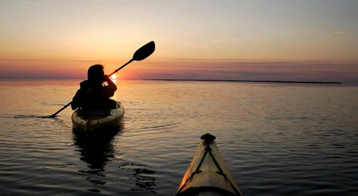 A peaceful scene of a kayaker gliding on calm water at sunset, with the glow of the setting sun reflecting off the water's surface. The image evokes a sense of adventure, tranquility, and connection to nature.